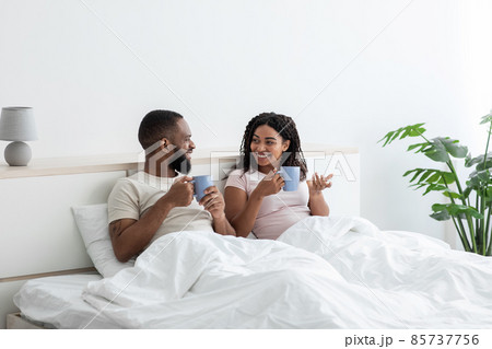 Cheerful young black husband and wife relaxing in bed with cups of coffee and talking, enjoy morning together Cheerful young black husband and wife relaxing in bed with cups of coffee and talking, enjoy morning together 85737756