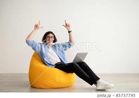 Happy freelance guy sitting in beanbag chair with brand new laptop, pointing up at copy space over light wall, panorama Happy freelance guy sitting in beanbag chair with brand new laptop, pointing up at copy space over light wall, panorama 85737795