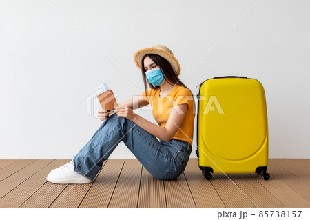 Young woman in face mask holding passport and tickets, sitting near suitcase over light wall background 85738157