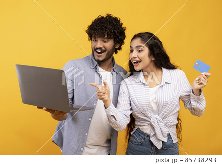 Joyful indian couple with laptop and credit card emotionally reacting to online profit, posing over yellow background Joyful indian couple with laptop and credit card emotionally reacting to online profit, posing over yellow background 85738293