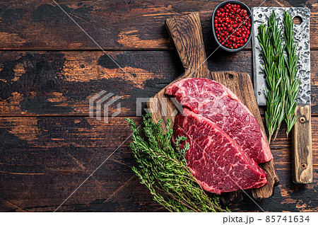 Raw denver cut black angus beef steak on a butcher board with meat cleaver. Dark wooden background. Top view. Copy space 85741634