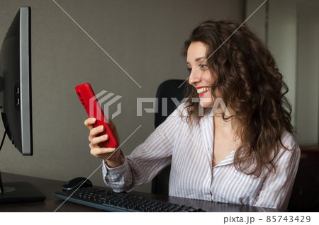 Tired young woman with curly hair and white shirt is working at the office and using her red smartphone, routine work, freelance, burnout syndrome 85743429