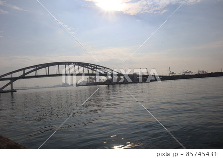 光り輝く太陽と青空と雲とアーチ橋と水面の光るところのある海とクレーンなどの港湾の風景 85745431