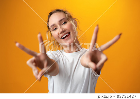 Portrait of a young funny woman showing peace gesture with two hands isolated over yellow background. 85751427