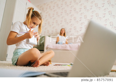 Low-angle view of cute primary girl child writing homework in copybook sitting on floor with laptop for distance education online at home. 85757091
