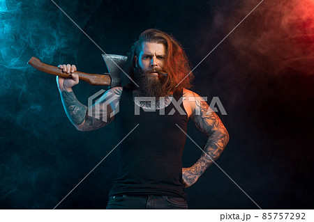 Handsome man with cigar holds old axe. Bearded lumberjack. Serious male with tattoos with long hair. Handsome man with cigar holds old axe. Bearded lumberjack. Serious male with tattoos with long hair. 85757292