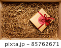 Wooden box with present wrapped in brown craft paper with red ribbon, filled with paper filler, top view 85762671