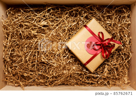 Wooden box with present wrapped in brown craft paper with red ribbon, filled with paper filler, top view 85762671