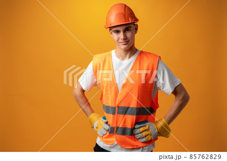 Confident smiling teen boy wearing orange hard hat against yellow background 85762829