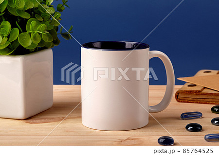 White ceramic mug on wooden desktop next to scattered glass stones, green plant in pot and brown notepad, blue background. Close up, copy space White ceramic mug on wooden desktop next to scattered glass stones, green plant in pot and brown notepad, blue background. Close up, copy space 85764525