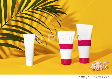 One white and two colorful cosmetic tubes with no logo next to a shell and green palm twig on yellow studio background. Close up, copy space One white and two colorful cosmetic tubes with no logo next to a shell and green palm twig on yellow studio background. Close up, copy space 85764557