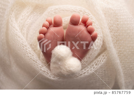 The tiny foot of a newborn baby. Soft feet of a new born in a white wool blanket. Close up of toes, heels and feet of a newborn. Knitted white heart in the legs of a baby. Macro photography.  85767759