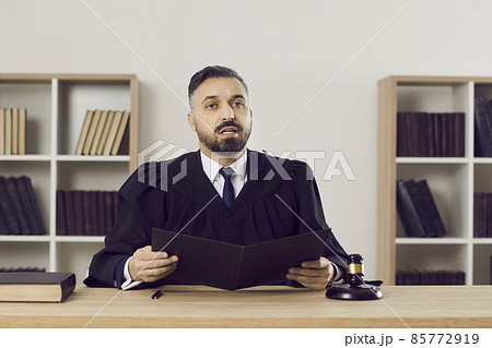 Judge passes judgement on the accused. Portrait of a young man in a robe reading the sentence while sitting at his table in the courtroom during a court session. Law, justice and legislation concept Judge passes judgement on the accused. Portrait of a young man in a robe reading the sentence while sitting at his table in the courtroom during a court session. Law, justice and legislation concept 85772919