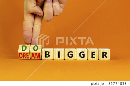 Dream and do bigger symbol. Businessman turns wooden cubes and changes words dream bigger to do bigger. Business and dream and do bigger concept. Beautiful orange background, copy space. 85774833