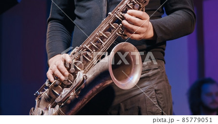 Man saxophonist at a concert plays the saxophone. Live musical performance and virtuosity of playing a brass musical instrument. It dark, colored spotlights are shining around Man saxophonist at a concert plays the saxophone. Live musical performance and virtuosity of playing a brass musical instrument. It dark, colored spotlights are shining around 85779051