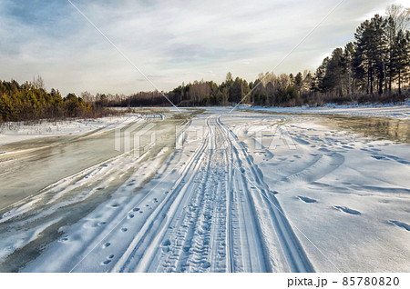 Winter landscape on a sunny day. Ski and snowmobile road along the river. Human footprints. The ice on the river has melted. Trees and bushes grow along the banks of the river. Beautiful sky  85780820