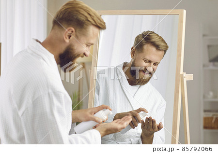 Smiling young man apply face cream for healthy smooth skin looking in mirror. Happy guy use balm do morning routine facial procedures at home. Male beauty concept. Skincare and treatment. 85792066