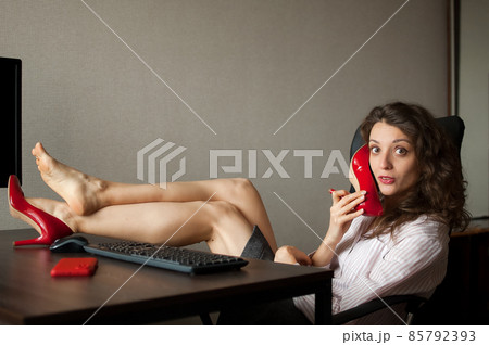 Crazy young woman is sitting at the desk with legs in red high heels on office table and pretending that she is talking by phone using one of her shoes Crazy young woman is sitting at the desk with legs in red high heels on office table and pretending that she is talking by phone using one of her shoes 85792393
