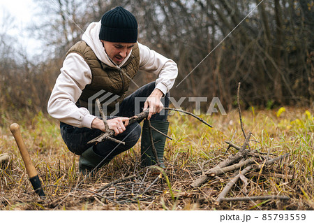 Portrait of frozen tourist man preparing firewood to campfire to keep warm and cook food at outdoors on overcast cold day. 85793559