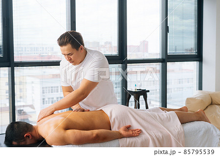 Side view of professional male masseur with strong tattooed hands massaging back and shoulder of muscular sports man lying on stomach Side view of professional male masseur with strong tattooed hands massaging back and shoulder of muscular sports man lying on stomach 85795539