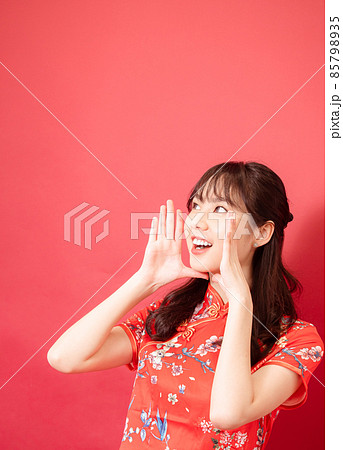 Young asian girl wear a red cheongsam is shouting and holding her hands near her mouth with copyspace on red background. Young asian girl wear a red cheongsam is shouting and holding her hands near her mouth with copyspace on red background. 85798935