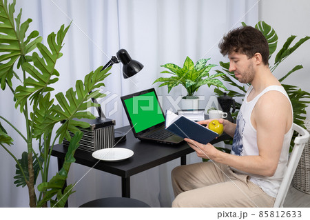 A young worker is holding a green apple in his hand while viewing a book calendar. A domain computer workstation among potted plants with bright lighting. Green screen of monitor ready to design. 85812633