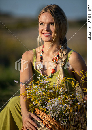 A young herbalist crouches by the dirt road with a basket full of herbs and looks into the sun. 85814676