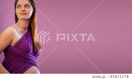 On a slightly pink background, there is a smiling young girl wearing a purple scarf in advanced pregnancy. Panoramic frame. 85815278