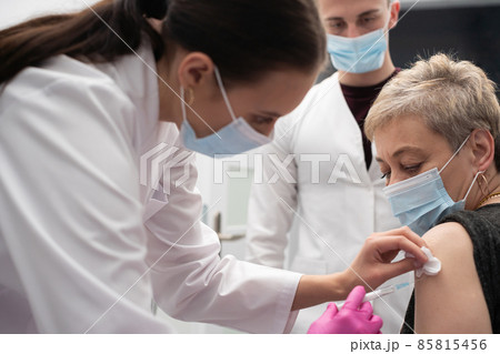 A nurse is making an vaccine to an elderly woman. The COVID vaccine19. A sterile doctor's office in a private clinic. A young medical intern looks at how to properly inject. 85815456
