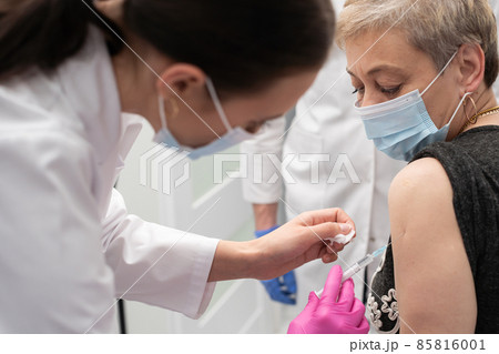 A nurse is making an vaccine to an elderly woman. The COVID vaccine19. A sterile doctor's office in a private clinic. A young medical intern looks at how to properly inject. 85816001