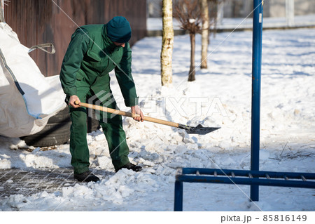 A manual worker scoops snow from the pavement and shovels it onto a pile next to it A manual worker scoops snow from the pavement and shovels it onto a pile next to it 85816149