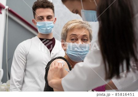 A nurse is making an vaccine to an elderly woman. The COVID vaccine19. A sterile doctor's office in a private clinic. A young medical intern looks at how to properly inject. A nurse is making an vaccine to an elderly woman. The COVID vaccine19. A sterile doctor's office in a private clinic. A young medical intern looks at how to properly inject. 85816451