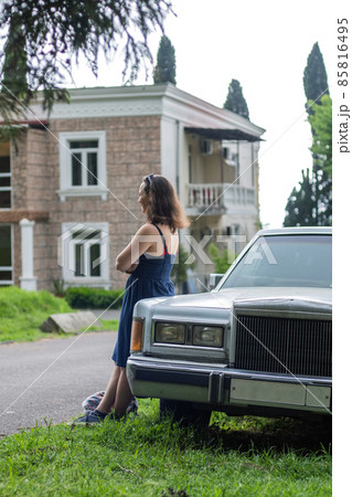 Young Woman Leaning on an Old Limo Car over Big Mansion in a Tropical Area 85816495