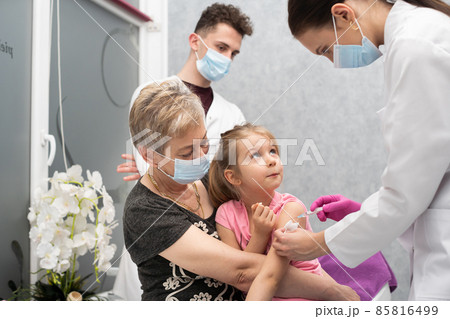 A nurse gives a vaccine to a girl who is sitting on her grandmother's lap because she feels so safe. A trainee in the last year of medicine observes the whole situation in the doctor's office and 85816499