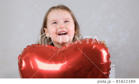 little cute girl in a red dress hides behind a red heart shaped balloon. valentines day concept 85822389