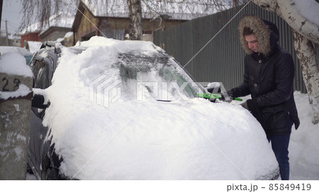 A young man in a jacket cleans his car of snow with a brush. The car was covered in snow. Weather disaster. 85849419