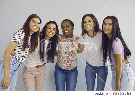 Multiracial female friends. Portrait of a group of happy different young women hugging and smiling sincerely together. Women in casual clothes look at the camera on a background of a light wall. 85855263