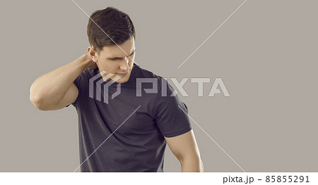 Back and spine disease. Young unhappy muscular man suffers from neckache due to muscle spasm or pinched nerve. Caucasian man in black T-shirt rubs his neck on gray background. Isolated. Banner. 85855291