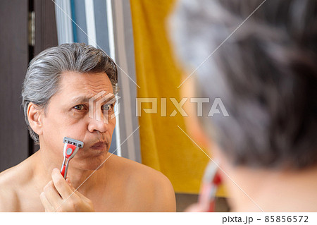 An adult gray-haired man shaves with a razor in front of a mirror 85856572
