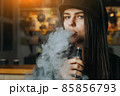 Young pretty woman in cap smoke an electronic cigarette at the vape shop. Hip-hop style. Closeup. 85856793