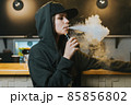 Young pretty woman in cap smoke an electronic cigarette at the vape shop. Hip-hop style. Closeup. 85856802