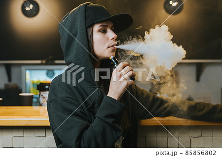 Young pretty woman in cap smoke an electronic cigarette at the vape shop. Hip-hop style. Closeup. Young pretty woman in cap smoke an electronic cigarette at the vape shop. Hip-hop style. Closeup. 85856802