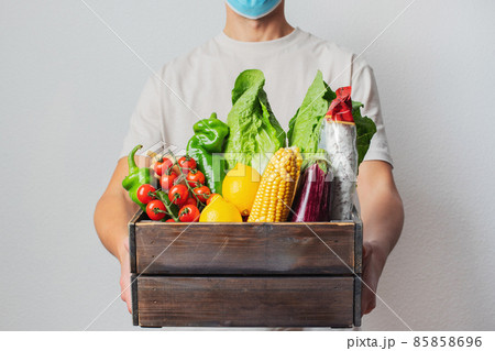 Young delivery man in protective face mask holding wooden box in hands. Man courier with shopping box. Safe online delivery from supermarket to home. COVID-19 coronavirus protection Young delivery man in protective face mask holding wooden box in hands. Man courier with shopping box. Safe online delivery from supermarket to home. COVID-19 coronavirus protection 85858696
