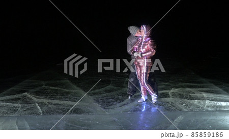 Young loving couple skating at ice rink at night. Man and woman kiss hug have fun learn to skate. Making love on ice. Ice illuminated backlit below. Young loving couple skating at ice rink at night. Man and woman kiss hug have fun learn to skate. Making love on ice. Ice illuminated backlit below. 85859186