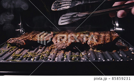 Delicious juicy meat steak cooking on grill. Aged prime rare roast grilling marble beef. Electric roaster, rosemary black pepper, salt. Kitchen tongs. Delicious juicy meat steak cooking on grill. Aged prime rare roast grilling marble beef. Electric roaster, rosemary black pepper, salt. Kitchen tongs. 85859231