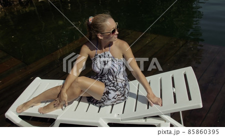 Woman lie on a sunbed in sunglasses and a boho silk shawl. Girl rest on a flood wood underwater pier. The pavement is covered with water in the lake. 85860395