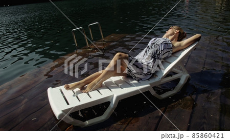 Woman lie on a sunbed in sunglasses and a boho silk shawl. Girl rest on a flood wood underwater pier. The pavement is covered with water in the lake. 85860421
