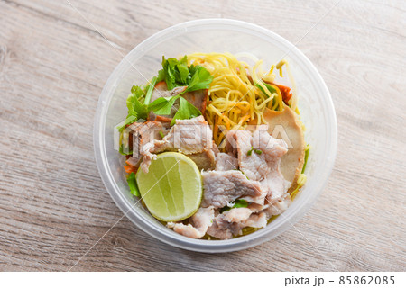 Dry noodles pork with red roasted pork and vegetable in plastic bowl on wooden table, top view Thai and China Asian food yellow noodles lemon lime 85862085