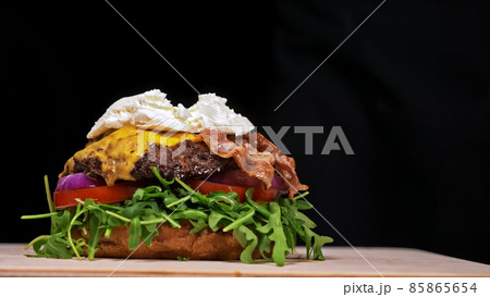 Craft burger is cooking on black background. Consist: sauce, arugula, tomato, onion, bacon, currant sauce, ricotta cheese, air bun and marble meat. 85865654