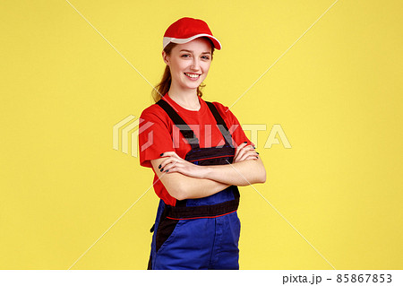 Portrait of adorable worker woman standing with crossed hands, looking at camera with positive confident expression, wearing overalls and red cap. Indoor studio shot isolated on yellow background. 85867853